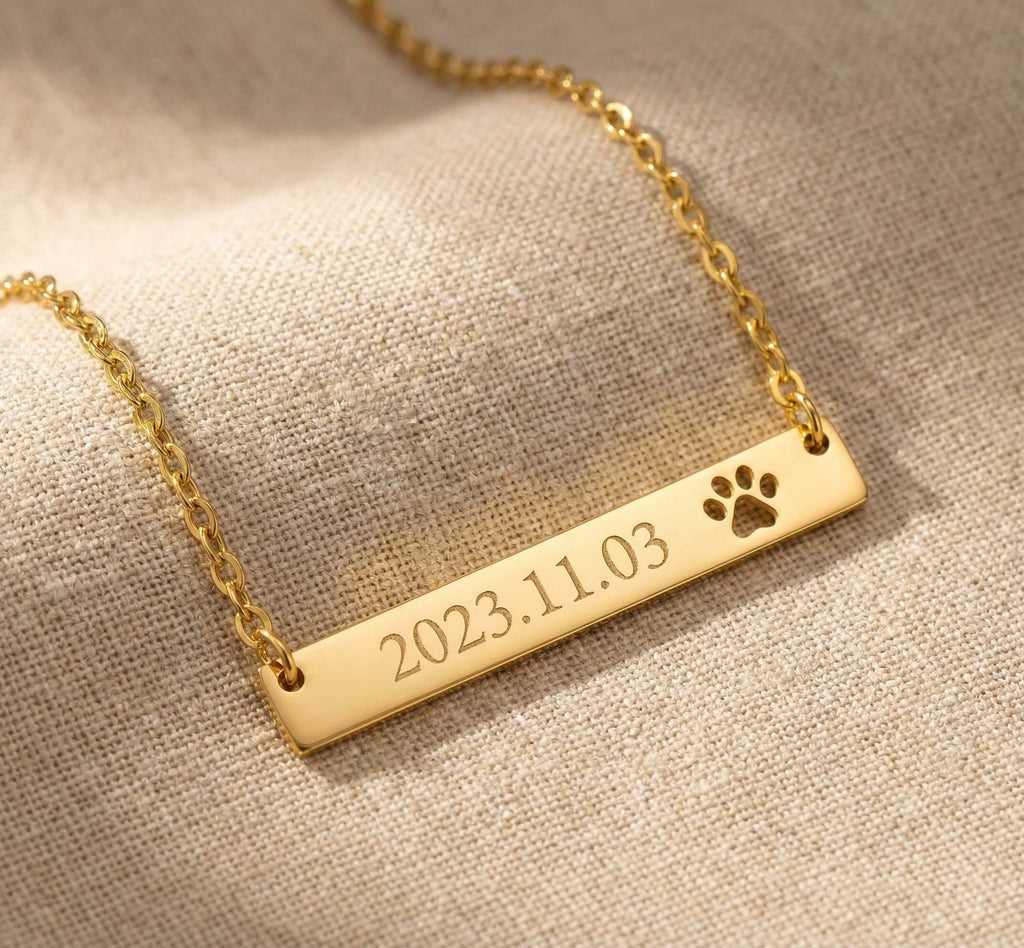 Gold necklace with a bar pendant featuring engraved date and paw print on a beige fabric background